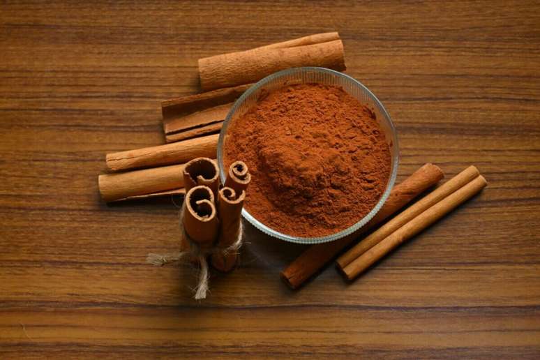 Uma composi&ccedil;&atilde;o sobre uma superf&iacute;cie de madeira marrom mostrando canela em duas formas: em p&oacute; e em pau. No centro, h&aacute; uma pequena tigela de vidro redonda cheia de canela em p&oacute; de cor marrom-avermelhada. Ao redor da tigela, est&atilde;o espalhados v&aacute;rios paus de canela secos. No canto inferior esquerdo, tr&ecirc;s paus de canela est&atilde;o amarrados verticalmente com um peda&ccedil;o de barbante r&uacute;stico.