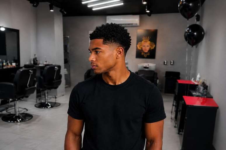 Homem com corte crop cacheado, usando blusa preta, com o rosto virado de lado
