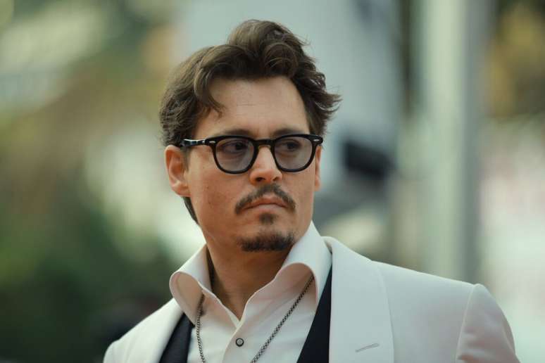 Fotografia em plano m&eacute;dio do ator Johnny Depp olhando de perfil parcial para a direita com uma express&atilde;o s&eacute;ria. Ele usa &oacute;culos de grau com arma&ccedil;&atilde;o preta, terno com palet&oacute; branco, colete preto e camisa social branca com o colarinho aberto.