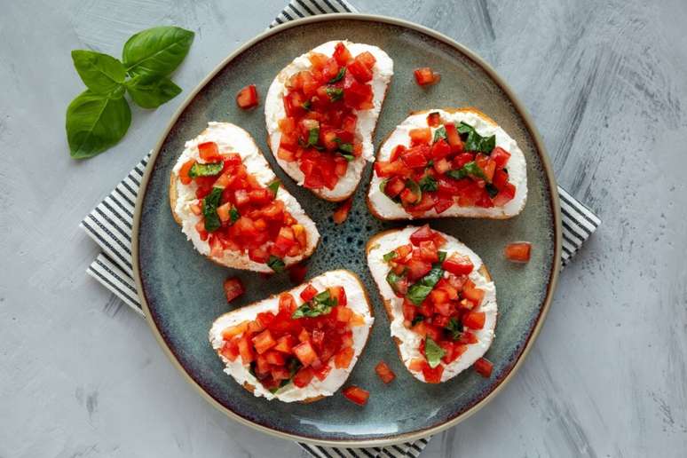 Bruschettas com ricota, tomate em cubos e folhas de manjeric&atilde;o servidas em fatias de p&atilde;o sobre um prato redondo