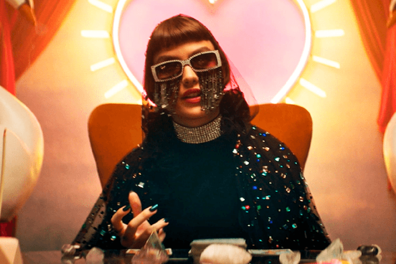 Mulher usando &oacute;culos decorados e roupas brilhantes, sentada diante de uma mesa com cristais sob uma ilumina&ccedil;&atilde;o colorida em formato de cora&ccedil;&atilde;o