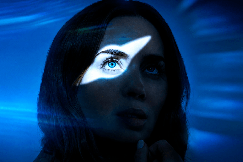 Mulher observando para cima enquanto um feixe de luz destaca um de seus olhos em um ambiente escuro e azulado
