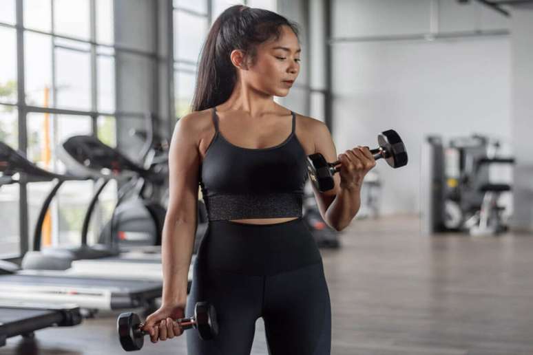 mulher de cabelo preso, regata e cal&ccedil;a pretas, segurando halteres nas m&atilde;os e flexionando os bra&ccedil;os