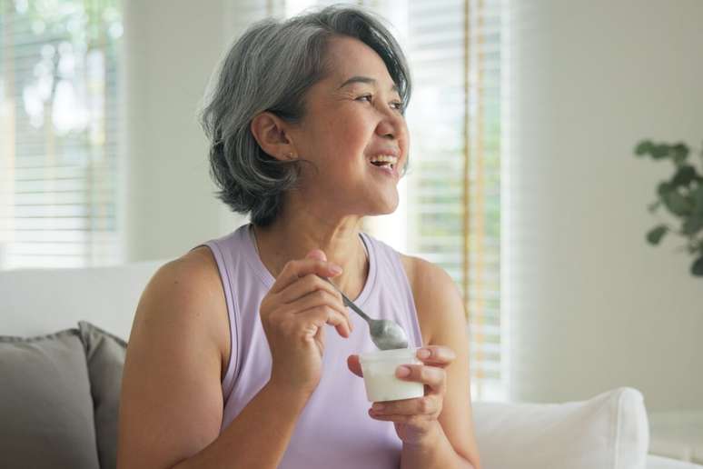 Uma mulher de meia-idade com tra&ccedil;os asi&aacute;ticos e cabelos curtos e grisalhos aparece sorrindo e olhando para o lado direito da imagem. Ela est&aacute; sentada em um sof&aacute; branco, veste uma blusa regata lil&aacute;s e segura um pote pequeno de iogurte branco em uma das m&atilde;os, enquanto usa uma colher com a outra m&atilde;o. O ambiente ao fundo &eacute; uma sala iluminada pela luz natural que entra atrav&eacute;s de uma janela com persianas.