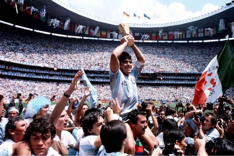 Uma imagem de corpo inteiro de Diego Maradona, um dos maiores jogadores de futebol de todos os tempos, levantando a Ta&ccedil;a da Copa do Mundo. Ele est&aacute; em cima dos ombros de um torcedor, com a multid&atilde;o e os jogadores da Sele&ccedil;&atilde;o Argentina ao seu redor. O fundo &eacute; um est&aacute;dio de futebol.