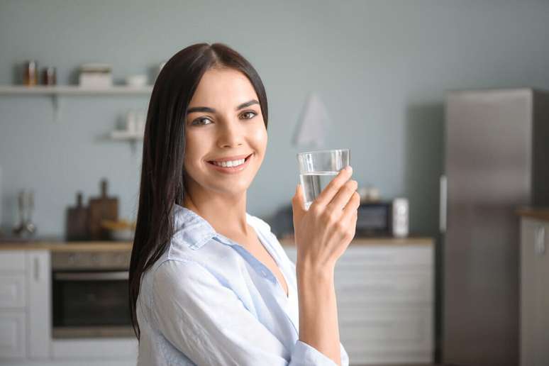 Mulher com o cabelo liso solto, usando camisa azul clara de mangar e com um copo de &aacute;gua na m&atilde;o, ao fundo est&aacute; a cozinha da casa