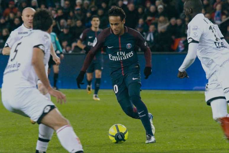 Uma imagem de corpo inteiro de Neymar Jr., um dos melhores jogadores de futebol da atualidade, com a camisa azul e vermelha do Paris Saint-Germain, o uniforme n&uacute;mero dez. Ele est&aacute; driblando com a bola, com dois jogadores advers&aacute;rios ao seu redor. O fundo &eacute; um campo de futebol.