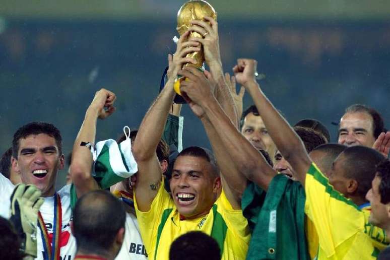 Uma imagem de corpo inteiro de Ronaldo Naz&aacute;rio, segurando a Ta&ccedil;a da Copa do Mundo. Ele est&aacute; sorrindo para a c&acirc;mera, com a medalha de campe&atilde;o ao redor do pesco&ccedil;o. O fundo &eacute; uma multid&atilde;o borrada em um est&aacute;dio.