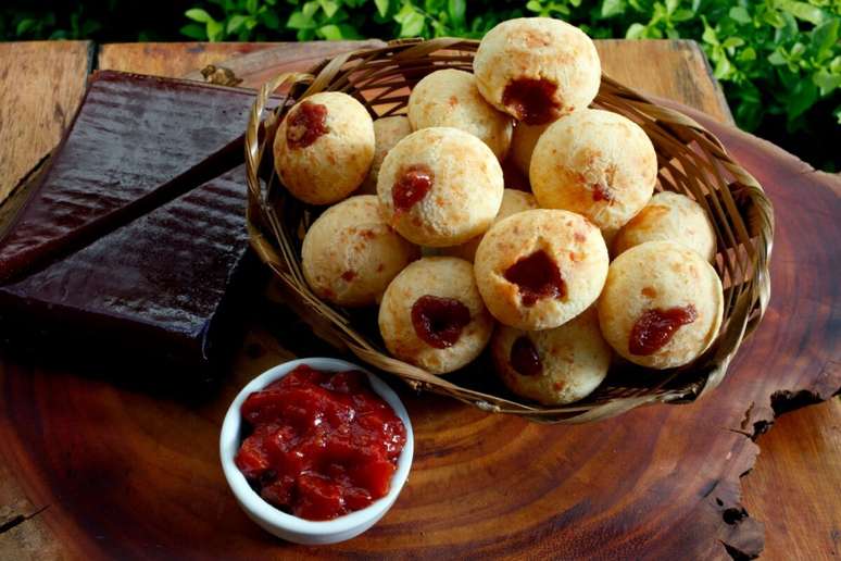 Uma cesta de vime cheia de p&atilde;es de queijo redondos, recheados com goiabada cremosa que aparece no topo de cada um. Ao lado, h&aacute; um bloco de goiabada casc&atilde;o e um pequeno pote com mais goiabada pastosa, dispostos sobre uma t&aacute;bua de madeira r&uacute;stica.
