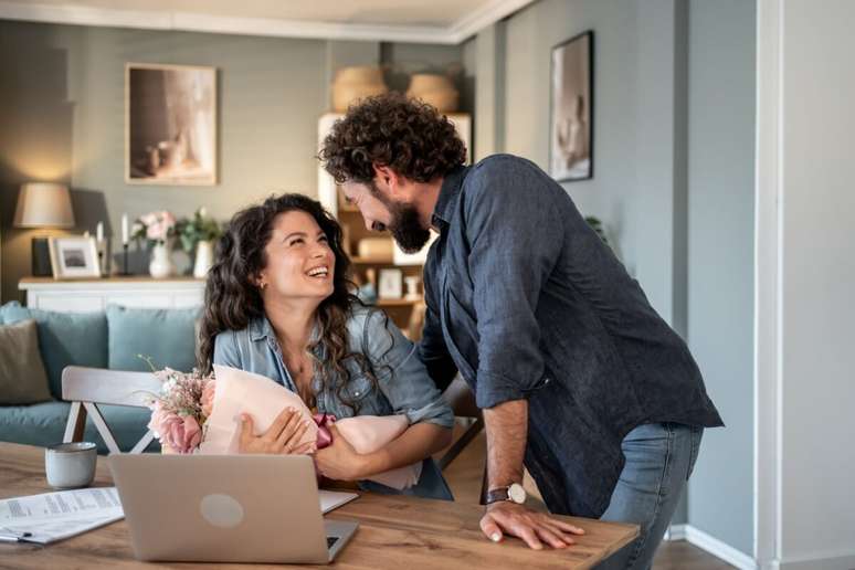 Homem com cabelo cacheado e barba entrega um buqu&ecirc; de flores para uma mulher sentada em frente a um notebook. Ambos sorriem e se olham com carinho em um ambiente residencial aconchegante.