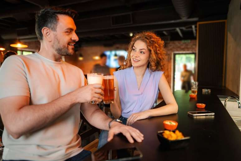 Homem e mulher sorriem e brindam com copos de cerveja no balc&atilde;o de um bar. O homem usa camiseta clara e a mulher, com cabelos cacheados, veste uma blusa lil&aacute;s sem mangas. Pequenas por&ccedil;&otilde;es de petiscos est&atilde;o espalhadas sobre o balc&atilde;o escuro.