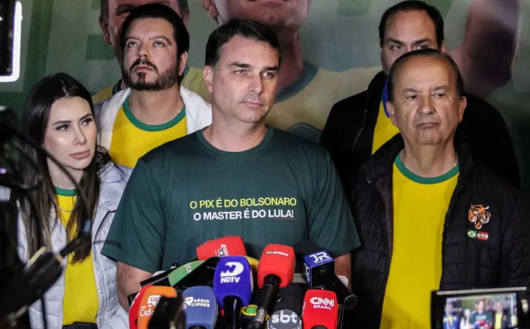 Fl&aacute;vio Bolsonaro cercado por apoiadores usando camisas amarelas enquanto Fl&aacute;vio usa uma verde escrito: "O pix &eacute; do Bolsonaro. O Master &eacute; do Lula"