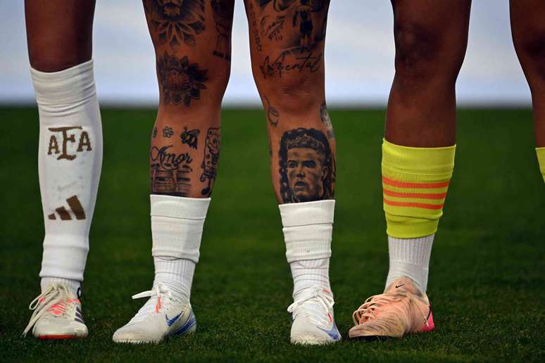 Pernas de quatro jogadores em um campo de futebol, duas delas cobertas de tatuagens.