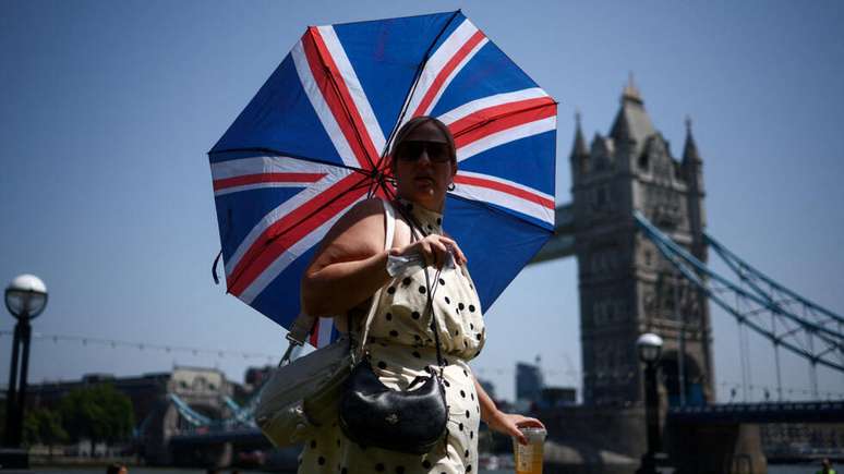 Uma mulher usa um guarda-chuva para se proteger do sol enquanto caminha perto da Tower Bridge, em Londres, Gr&atilde;-Bretanha, em 25 de junho de 2026, em meio a temperaturas recordes que afetam escolas e o transporte p&uacute;blico.