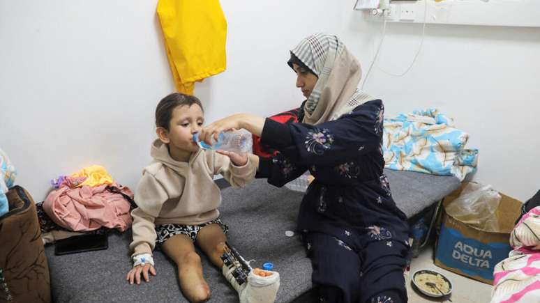 A menina palestina Eman Al-Kholi, que teve um membro amputado ap&oacute;s ser ferida em um ataque israelense que matou seus pais, recebe ajuda de um parente para beber &aacute;gua enquanto &eacute; tratada no Hospital Europeu, em Rafah, no sul da Faixa de Gaza, em 28 de dezembro de 2023.