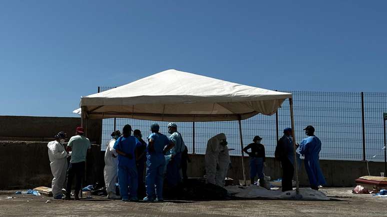 Equipes m&eacute;dicas e da per&iacute;cia trabalham ao lado de corpos cobertos por sacos pl&aacute;sticos e estendidos no ch&atilde;o