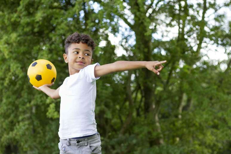 Uma crian&ccedil;a segura uma bola de futebol atr&aacute;s da cabe&ccedil;a, pronta para arremessar, enquanto aponta para a frente com a outra m&atilde;o.