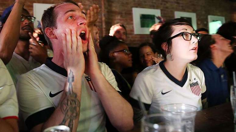 Dois torcedores de futebol dos Estados Unidos reagem durante a partida entre Gana e EUA no bar WoodWork, no bairro do Brooklyn, em Nova York. Um &eacute; homem e a outra &eacute; mulher. Ambos vestem camisetas da sele&ccedil;&atilde;o americana de futebol.