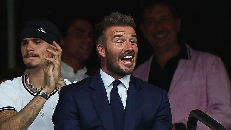 O ex-jogador de futebol ingl&ecirc;s David Beckham sorri durante a partida das quartas de final da Copa do Mundo de 2026 entre Noruega e Inglaterra, no Est&aacute;dio de Miami, em Miami Gardens, no dia 11 de julho. Ele veste um terno azul, camisa branca e gravata azul.