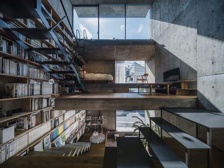 Casa com paredes de concreto, v&aacute;rias escadas e uma estante de livros que ocupa toda a parede esquerda