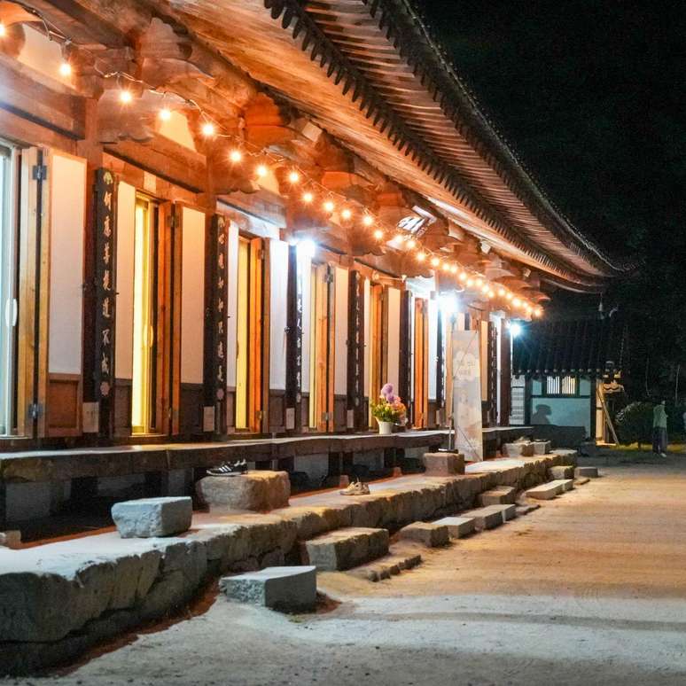 Templo coreano tradicional iluminado por luzes acolhedoras &agrave; noite, com um telhado curvo, colunas de madeira, escadas de pedra rodeando um amplo p&aacute;tio e visitantes caminhando pelo corredor que rodeia o templo