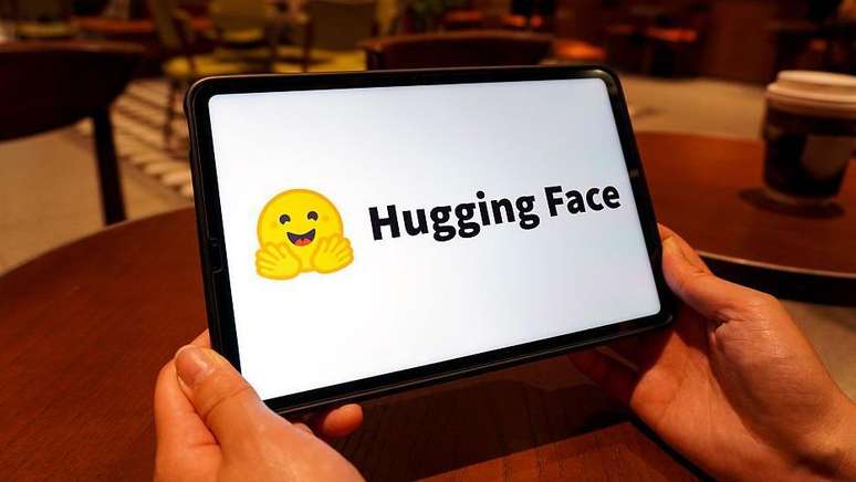O logotipo do Hugging Face &eacute; exibido na tela de um tablet. &Eacute; um emoji amarelo de um rosto sorridente com as m&atilde;os abertas &agrave; frente.