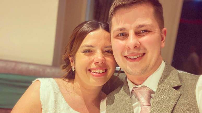 Uma foto de casamento de Josh e Kelly. Kelly usa um vestido de noiva branco, sem mangas e bordado, e brincos de p&eacute;rola; seu cabelo est&aacute; preso em um penteado para cima. Josh veste uma camisa branca, gravata bordada em rosa-claro e colete bege. Ambos olham para a c&acirc;mera e sorriem.