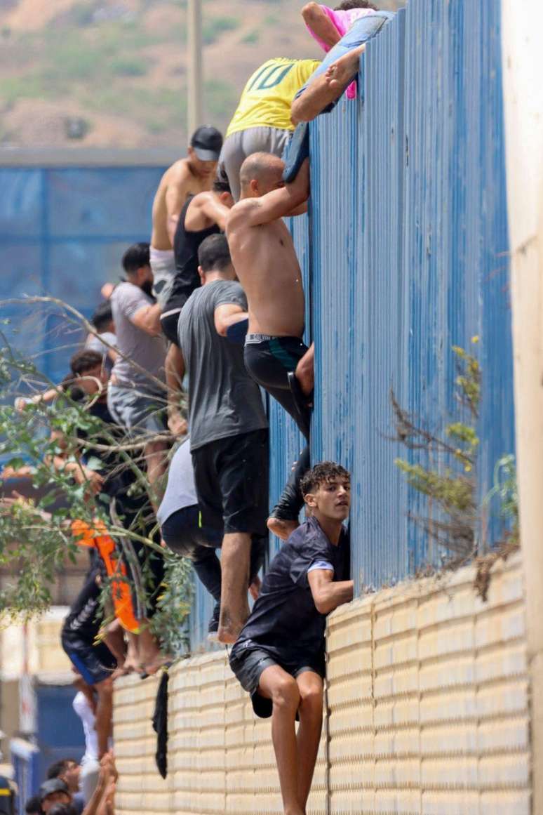 Migrantes escalam uma cerca em Ceuta, territ&oacute;rio espanhol no norte da &Aacute;frica, ap&oacute;s atravessarem a nado a partir do Marrocos, em 30 de julho de 2026.