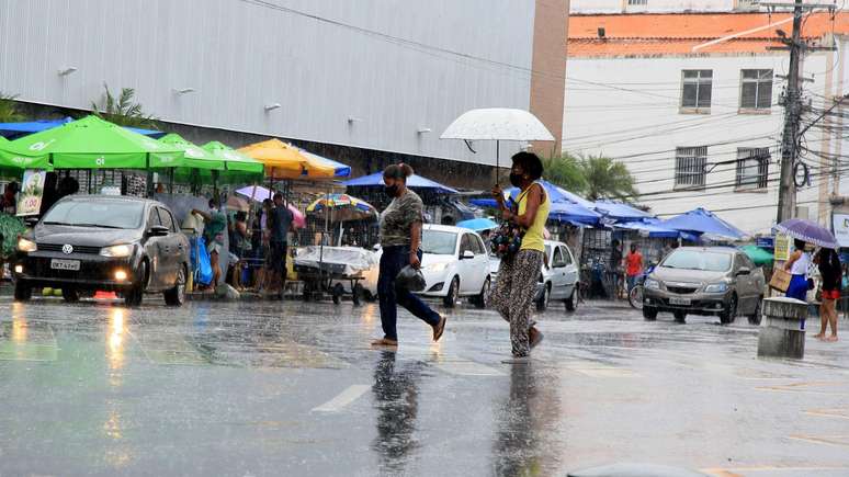 Chuva em rua movimentada no Brasil. Mulheres andam em faixa de pedestres, uma delas usa um guarda-chuva.