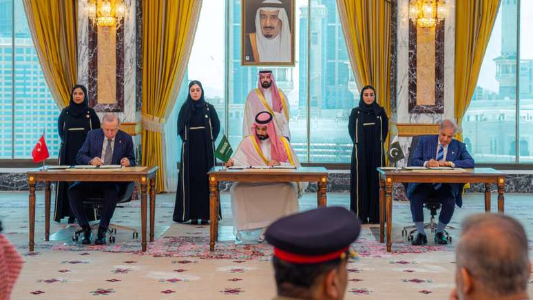 O presidente turco, Recep Tayyip Erdogan, o pr&iacute;ncipe herdeiro saudita, Mohammed bin Salman, e o primeiro-ministro paquistan&ecirc;s, Shehbaz Sharif, assinam acordo de defesa m&uacute;tua em Meca, na Ar&aacute;bia Saudita, em 7 de agosto de 2026. O pacto foi firmado em meio &agrave; escalada das tens&otilde;es no Oriente M&eacute;dio.