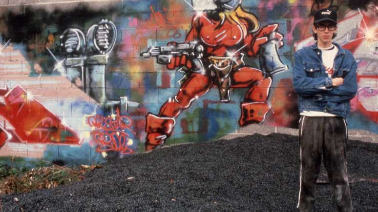 Fotografia de Henry Chalfant mostra o artista franc&ecirc;s Bando diante de uma obra de Mode 2, um dos principais nomes da arte urbana europeia. A imagem documenta o interc&acirc;mbio entre artistas que ajudaram a difundir a cultura do grafite para al&eacute;m de Nova York, consolidando sua presen&ccedil;a na cena art&iacute;stica internacional.