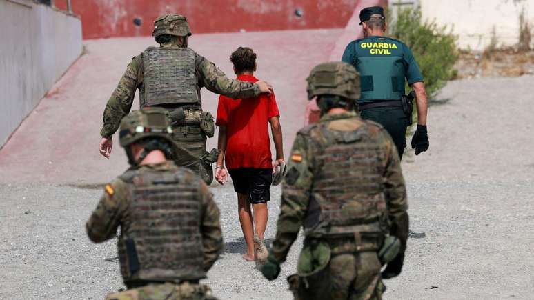 Tr&ecirc;s soldados e um agente da Guarda Civil acompanham um menor vestindo uma camiseta vermelha e bermuda. Todos est&atilde;o de costas. Ceuta, 1&ordm; de agosto de 2026.