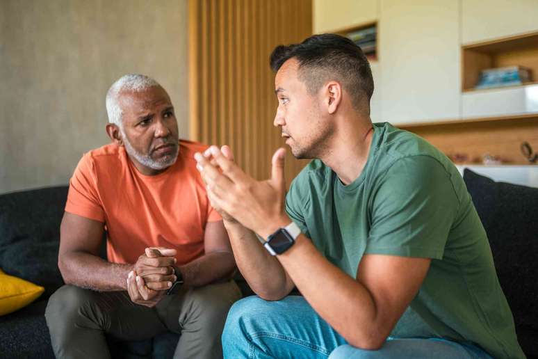 Dois homens de meia-idade, de diferentes origens raciais, conversam intimamente sentados em uma sala de estar.
