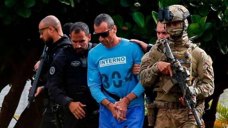 Detento algemado e com uniforme azul &eacute; escoltado por agentes de seguran&ccedil;a fortemente armados