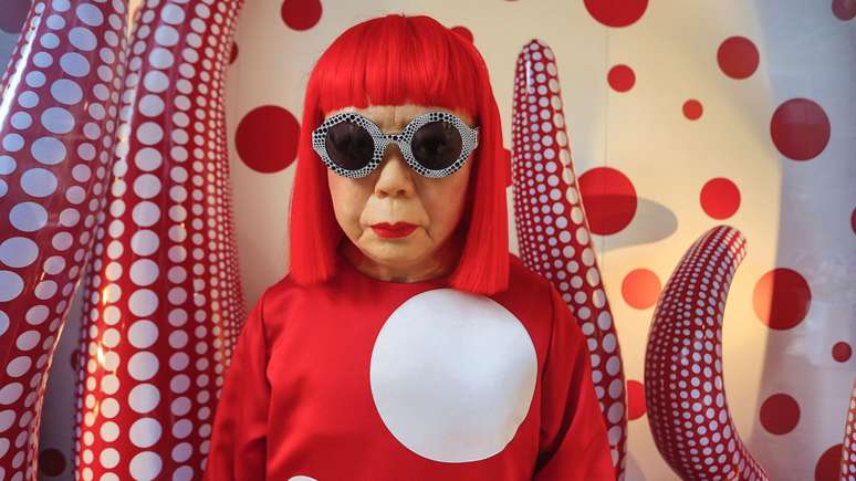 Yayoi Kusama com peruca vermelha, blusa vermelha com um c&iacute;rculo gigante e &oacute;culos, em frente a uma obra de arte vermelha e branca com c&iacute;rculos, que lembra um tent&aacute;culo