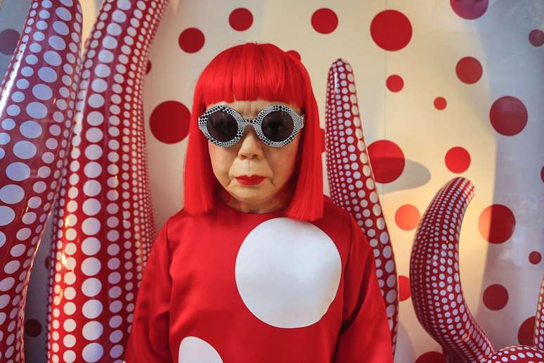 Yayoi Kusama com peruca vermelha, blusa vermelha com um c&iacute;rculo gigante e &oacute;culos, em frente a uma obra de arte vermelha e branca com c&iacute;rculos, que lembra um tent&aacute;culo