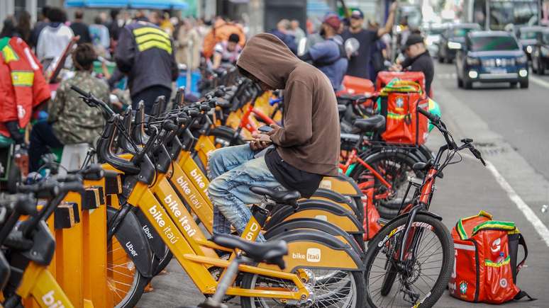 Jovem sentado em uma das dezenas de bicicletas por aluguel do Ita&uacute;, olhando para o celular