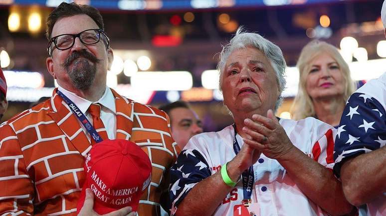 Um homem vestido com um terno na cor de tijolo est&aacute; de p&eacute; durante a conven&ccedil;&atilde;o. Ele segura um bon&eacute; com a frase &ldquo;Make America Great Again&rdquo;. Ao lado dele, uma mulher est&aacute; de p&eacute;, aplaudindo