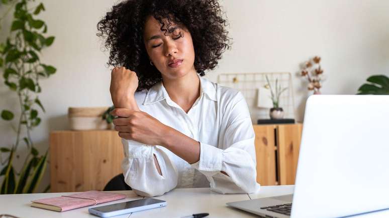 Mulher em frente a um laptop e um telefone celular, segurando o pulso com express&atilde;o de dor