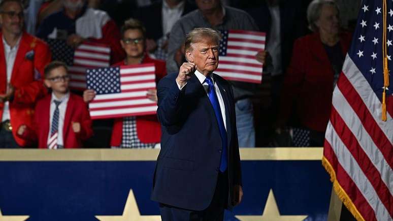 O presidente Trump, vestindo um terno azul e gravata, levanta o punho no palco. Atr&aacute;s dele, h&aacute; uma multid&atilde;o segurando cartazes com a bandeira americana.