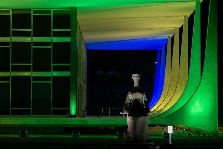 Foto noturna do STF, com est&aacute;tua da Justi&ccedil;a na frente e colunas em cores da bandeira brasileira 