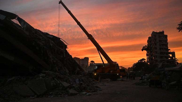 Entardecer com uma grua gigante removendo escombros em meio a estruturas que desabaram
