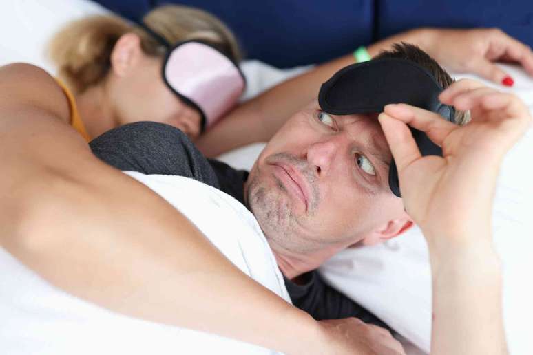 homem na cama levanta a m&aacute;scara para os olhos e olha intrigado para uma mulher adormecida com a m&aacute;scara