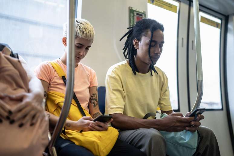 Uma mulher e um homem sentados num vag&atilde;o de metro olham para a tela do celular 