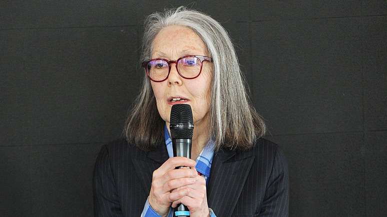 Anne Carson, mulher de cabelos grisalhos e &oacute;culos, segurando um microfone durante uma apresenta&ccedil;&atilde;o.