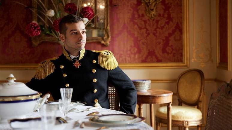 Jonas Lauwiner, sentado &agrave; mesa e vestindo uniforme militar, em um dos sal&otilde;es de seu "pal&aacute;cio". Sobre a mesa h&aacute; pratos e porcelana fina, enquanto as paredes est&atilde;o cobertas por tapetes vermelhos e dourados.