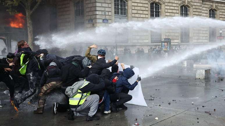 Manifestantes buscam prote&ccedil;&atilde;o atr&aacute;s de uma barricada enquanto a tropa de choque francesa utiliza canh&otilde;es de &aacute;gua em meio a confrontos durante uma manifesta&ccedil;&atilde;o na Place de la R&eacute;publique, em Paris