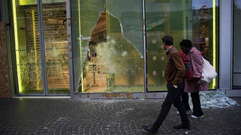 Pessoas passam, na manha de 19 de outubro de 2026, diante de vitrine de loja vandalizada durante protestos no centro de Paris, em 17 de outubro de 2026.