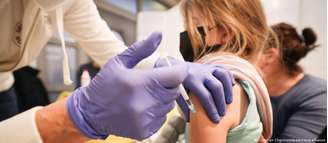 Mãos com luvas azuis aplicam vacina em uma menina. Ela segura um bicho de pelúcia. Não é possível ver o rosto dela. 