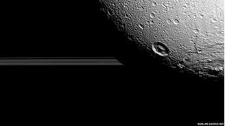 A lua Dione fotografada pela sonda Cassini com os anéis de Saturno ao fundo